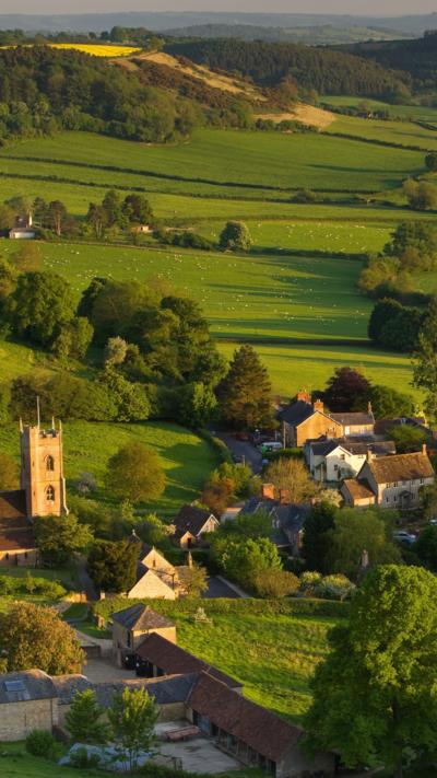 An aerial view of the countryside in Somerset with a small village with a church tower in the foreground and green hill and hedgerows and trees continuing into the distance