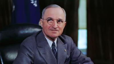A colour photo of US President Harry Truman wearing a grey suit, tie and glasses and smiling towards the camera. He is sat on a black leather chair behind a wooden desk while holding a yellow pencil above a notepad, as if he is about to write something on the paper. There are windows with curtains and a blue flag draped behind him.