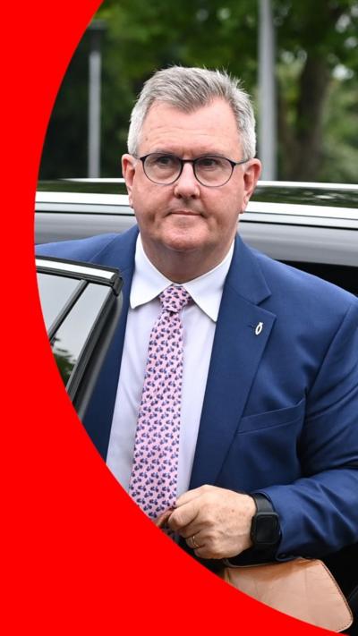 Jeffrey Donaldson walks into court wearing a blue suit, a white shirt and a pink patterned tie. He is holding a brown satchel. He is also wearing a pair of glasses. He has short, grey hair.