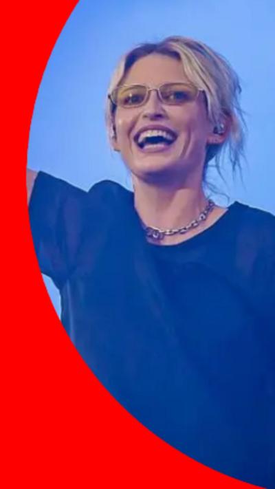 Emily Armstrong has short blonde hair and is wearing yellow glasses and a navy blue t-shirt on stage.