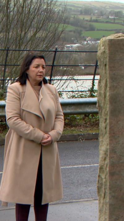 A woman in a camel-coloured coat stands with her hands clasped, in front of a large stone. It's not visible what's on the stone, but it could be a gravestone or memorial