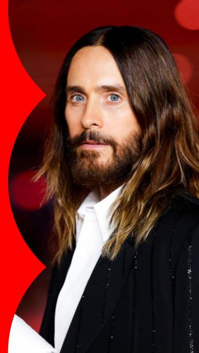 Jared Leto wears a black jacket and white shirt against a red background with a neutral expression