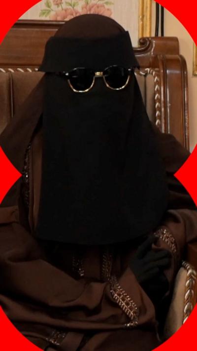A Muslim woman sits in a chair. She is wearing a headscarf and full face veil, with glasses.