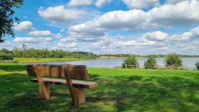 A wooden bench in a parkland area facing a lake under a blue sky with broken white clouds