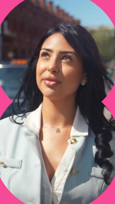 A woman with long black hair, in a light blue suit, looks up while walking in the street