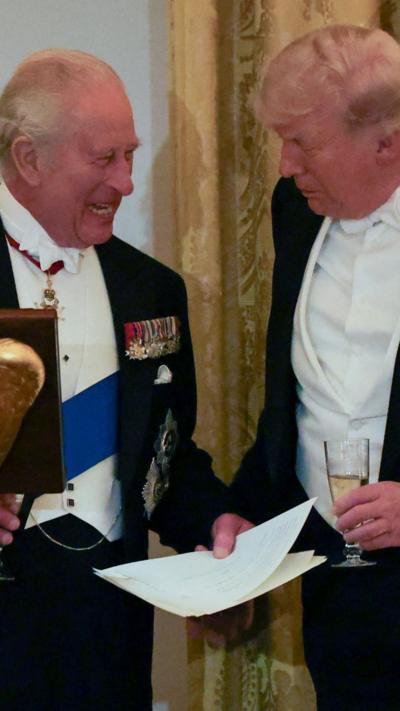 King Charles and President Trump stand together holding wine glasses, in formal dress
