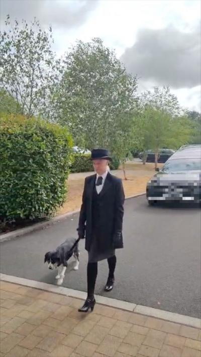 A woman in a black suit with a white shirt, black shoes, gloves and black bowler hat is walking in front of a hearse with a dog 