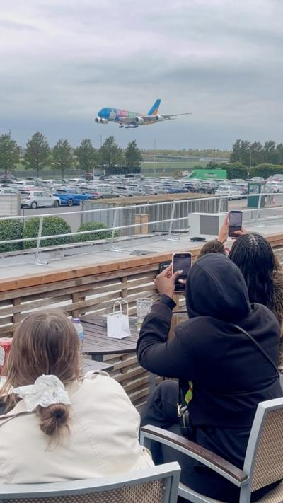 A colourful airplane descends towards an airport while onlookers take photos from a nearby observation area.