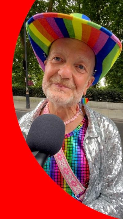 Keith, 64, wearing a rainbow cowboy hat, rainbow vest and silver sequinned jacket, speaks into a microphone.