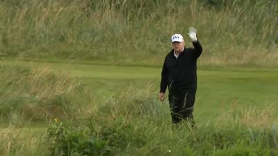 Man in a white baseball cap waves while standing on a golf course.