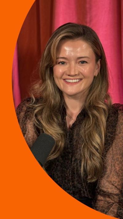 A woman with long brown hair, in front of a pink curtain, smiling to camera.