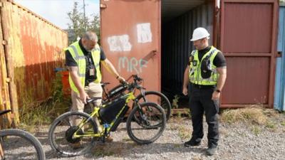Two men outside a container. They are both wearing hi-vis. One is holding an illegal e-bike.