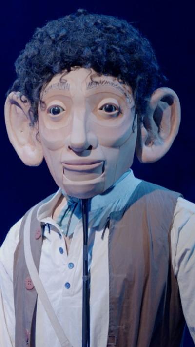 A puppet of a giant with black hair, big ears, grey shirt and brown waistcoat