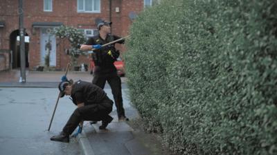 Two uniformed police officers search on a residential street. There is a large bush on the right hand side