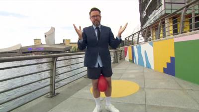 Jon Kay, in shorts has a red balloon between his knees, dressed from the waist up in a suit, shirt and tie