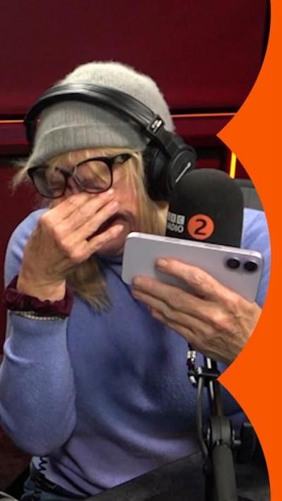 A blonde woman wearing a hat, puts her fingers over her eyes in laughter, in front of a Radio 2 mic.