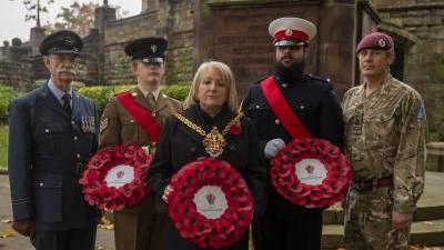 Mayor of Wolverhampton Linda Leach stands at the centre of a line, with two people on each side of her wearing military attire. She is wearing a black coat and her gold chains of office, whilst holding a poppy wreath. Two of the military personnel either side of her are also holding wreaths.