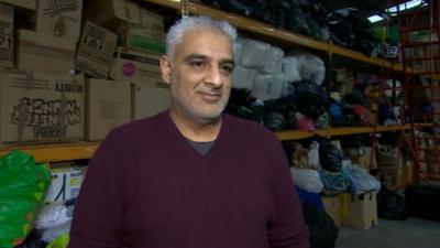 Tariq Jahan in front of piles of clothing and bedding donated by the public for migrants and refugees arriving in Greece