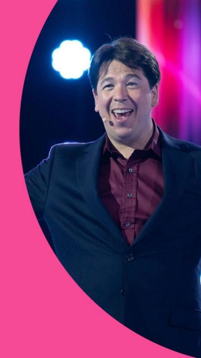 A man in a dark suit with a purple shift smiles. He has a Madonna mic on