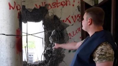 A wall with a hole blasted through it, a man is pointing at it