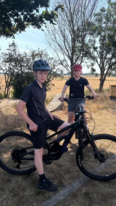 Two teenage boys, both wearing helmets, sit on mountain bikes in a wooded area. Behind them is a wooden bike ramp. 