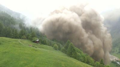 A large cloud generated by rock and mud rises over the green slope of a valley