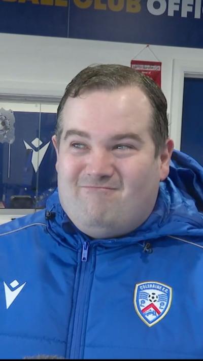 Jonathan Fillis is a man with dark short hair. He is standing in the shop. He is wearing a bright blue sports coat with the Coleraine FC emblem on the right hand side.