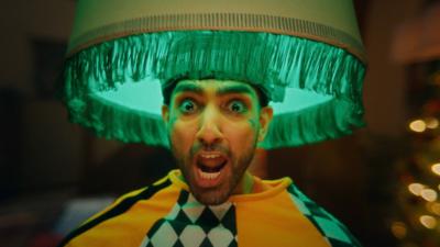 A man with his mouth open, looking shocked. He has a lampshade on his head and has on a baggy orange, black and white top