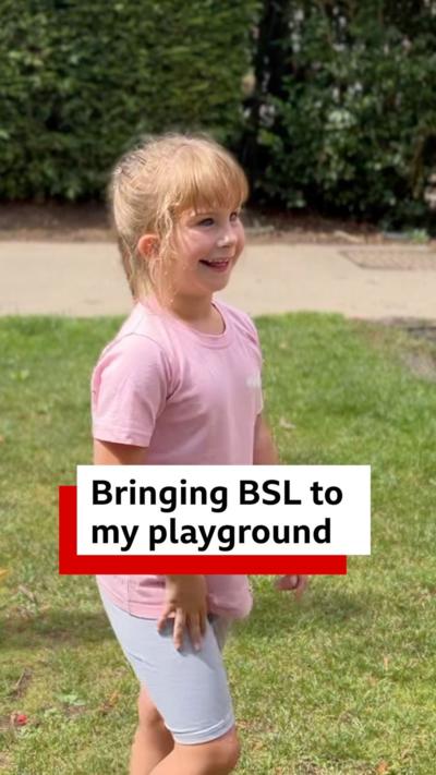 Emily is wearing a pink T shirt and is pictured in a park with lawns and hedges.
