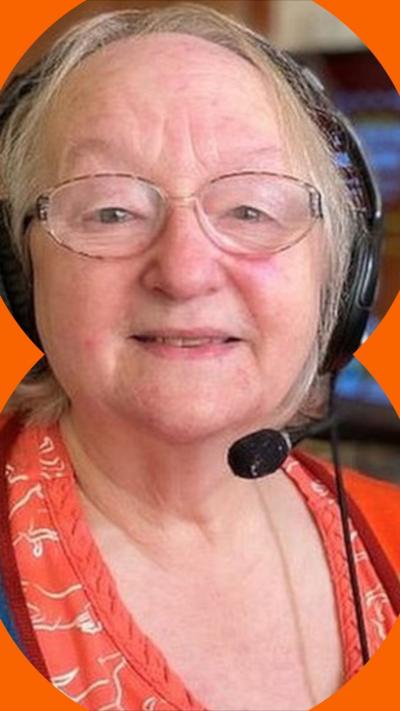 An elderly woman wears a gaming headset. There is a TV screen behind her 