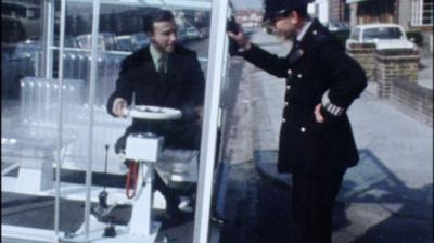 Presenter on left of frame sitting behind the wheel of the windowed city car.  He is clearly visible in the windowed box and the passenger seat and bench behind is also translucent.  A policeman on the right hand side is leaning against the side of the windowed car.