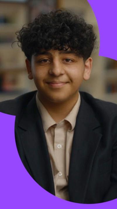 A teenage boy with black curly hair, wearing a beige shirt and navy blazer, smiling to camera. Blurred out bookshelves are behind.