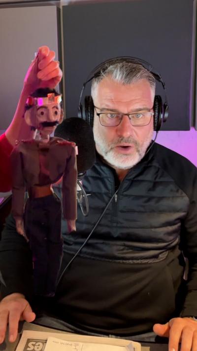 A man wearing glasses, headphones and a black jacket looks directly at the camera with a surprised expression. In front of him is a hand holding a puppet