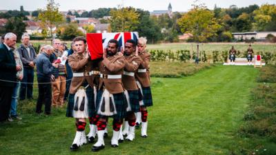 Six soldiers from the Royal Regiment of Scotland in kilts carry a coffin draped in the union flag at a cemetery in France.