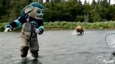 Someone dressed as Buoy the NHL mascot and wearing high waders, is seen running in knee deep water while being chased by a bear. Another man wearing fishing gear is also fleeing the running animal. In the background is a forest. 