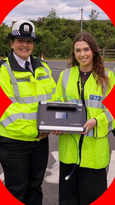 Two women, one in Police uniform and the other in a Police high-viz hold a breathalyser and smile at the camera.