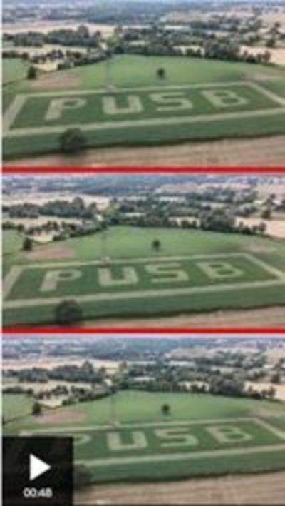 Aerial view of countryside with trees and hedges and green and yellow fields. The letters P U S B are cut into the nearest field.