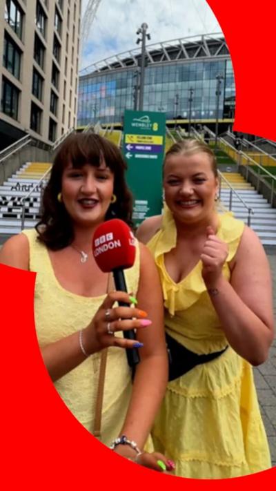 Two women in yellow dresses stand in front of Wembley Stadium smiling