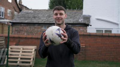Man holding football looking at the camera with a wall and fence behind him.