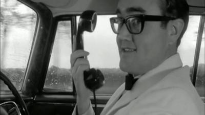 James Burke holding a telephone receiver in a car