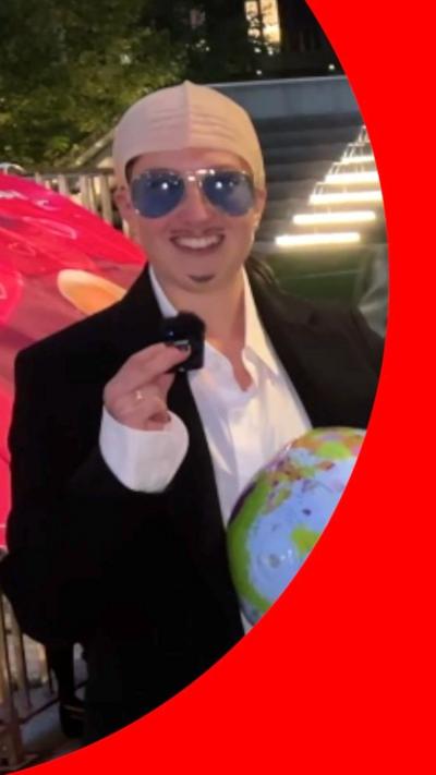 Woman dressed as Pitbull in sunglasses, bald cap and black blazer.
