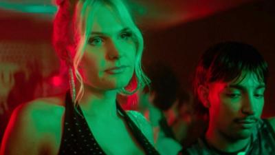 Sasha is wearing a black top. She is in a bar. Green and red lights are shining on her.