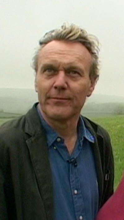 An old picture of Anthony Head pictured outside. A field can be seen behind him. He has short fair hair that's blown back by the wind. He is wearing a blue top and dark jacket.