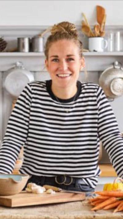 A woman, Helen Upshall, wears a striped jumper and has her hair in a messy bun. She stands at a worktop with cooking equipment in front of her.