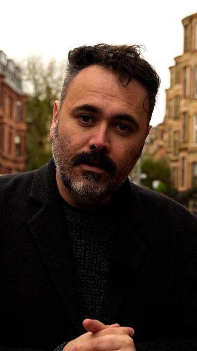 A man with short dark hair and a greying beard standing in a Glasgow tenement street.