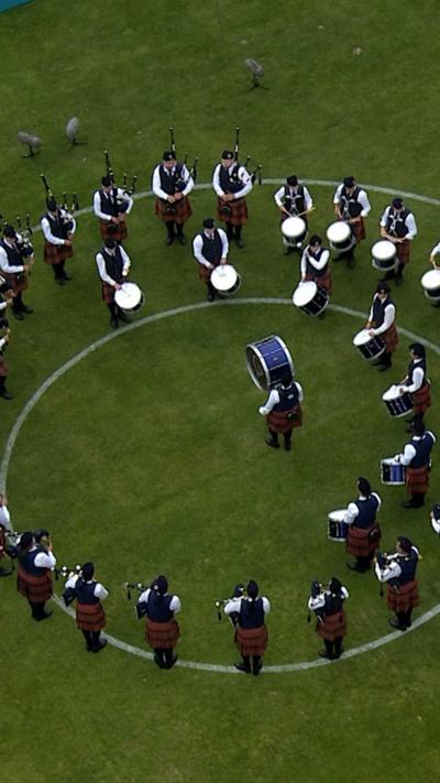 Large pipe band playing instruments in a circle formation, standing in a grassed area
