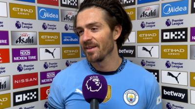 Jack Grealish in Man City kit in front of microphone.