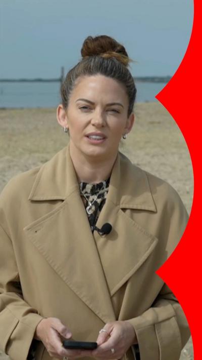A woman with with brown tied-up hair wearing a brown coat stands on beach speaking to the camera