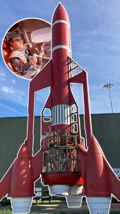 A 60ft tall red, Thunderbird rocket. It's on a grass field in front of a green building. It has white accent stripes