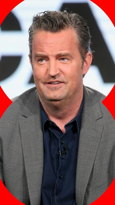 Matthew Perry has gelled back dark grey hair. He is wearing navy shirt and a grey suit jacket.